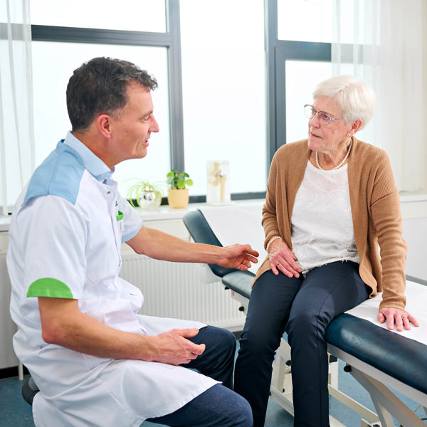Patiënt zit op behandeltafel, naast haar zit een dokter op een stoel. ze zijn met elkaar in gesprek. THEMA vormt de 'linking pin' tussen de Anna Zorggroep en haar zorgpartners