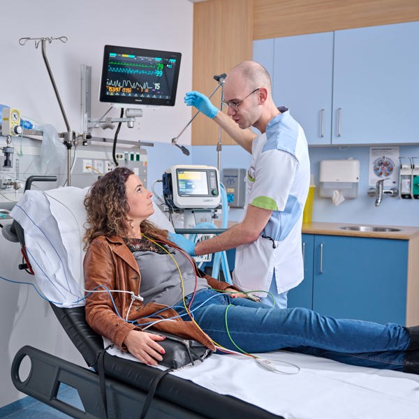 Patiënte ligt met jas aan op behandeltafel bij de spoedeisende hulp. Ze wordt gemonitord via apparatuur. Een zorgverleners staat naast haar; ze zijn in gesprek. THEMA vormt de 'linking pin' tussen de Anna Zorggroep en haar zorgpartners