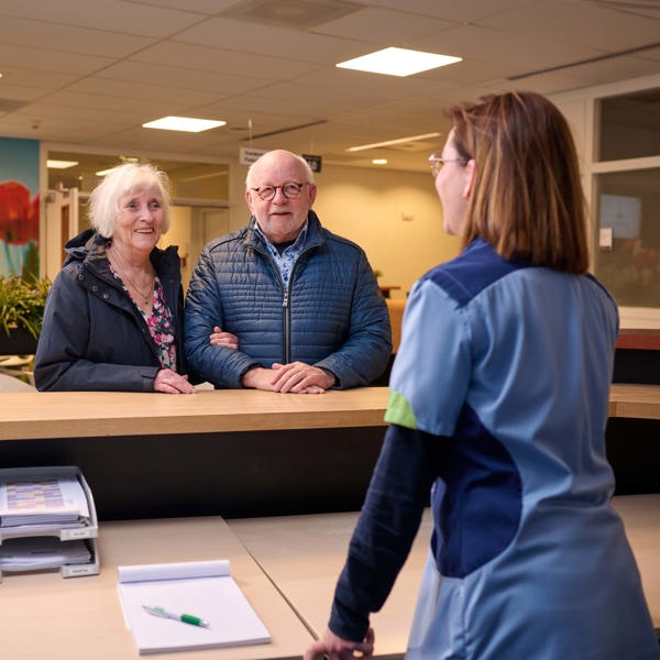 Mevrouw en meneer staan aan een balie, ze zijn in gesprek met zorgverlener aan de andere kant van de balie THEMA vormt de 'linking pin' tussen de Anna Zorggroep en haar zorgpartners